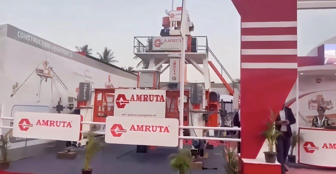 Amruta branded railings and logo boards around construction machinery beside the red arch column