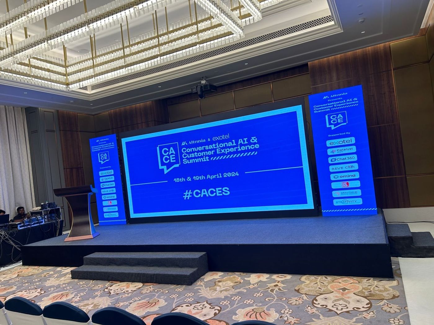 CACES summit stage in a hotel ballroom with a wide LED screen, two tall sponsor screens and carpeted platform