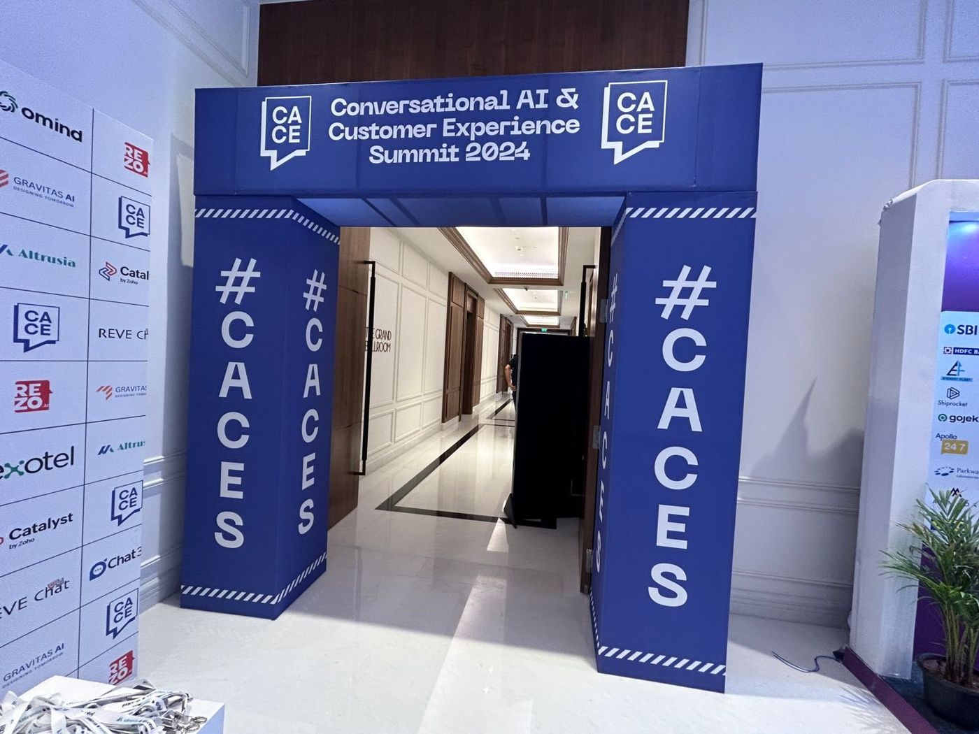 Blue CACES entry arch with summit title header and #CACES pillars beside the sponsor logo wall
