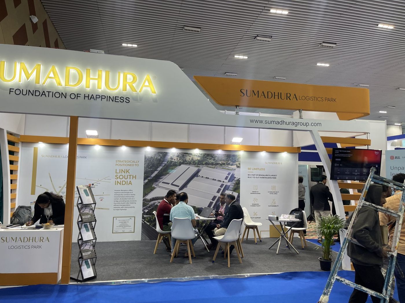 Sumadhura corner stall with backlit yellow lettering, curved mustard banner and open meeting lounge