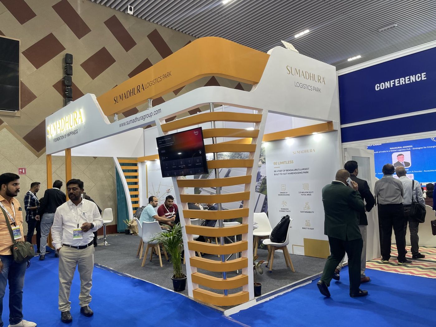 Corner view of the Sumadhura stall showing the slatted mustard tower and curved raised banner