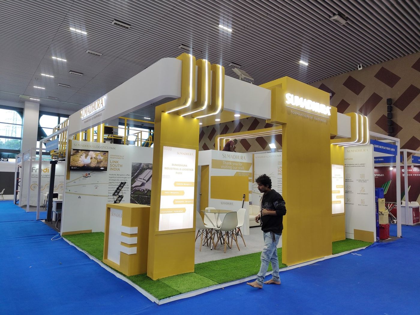 Sumadhura stall with mustard portals, white fascia, LED strip lines and artificial grass edging