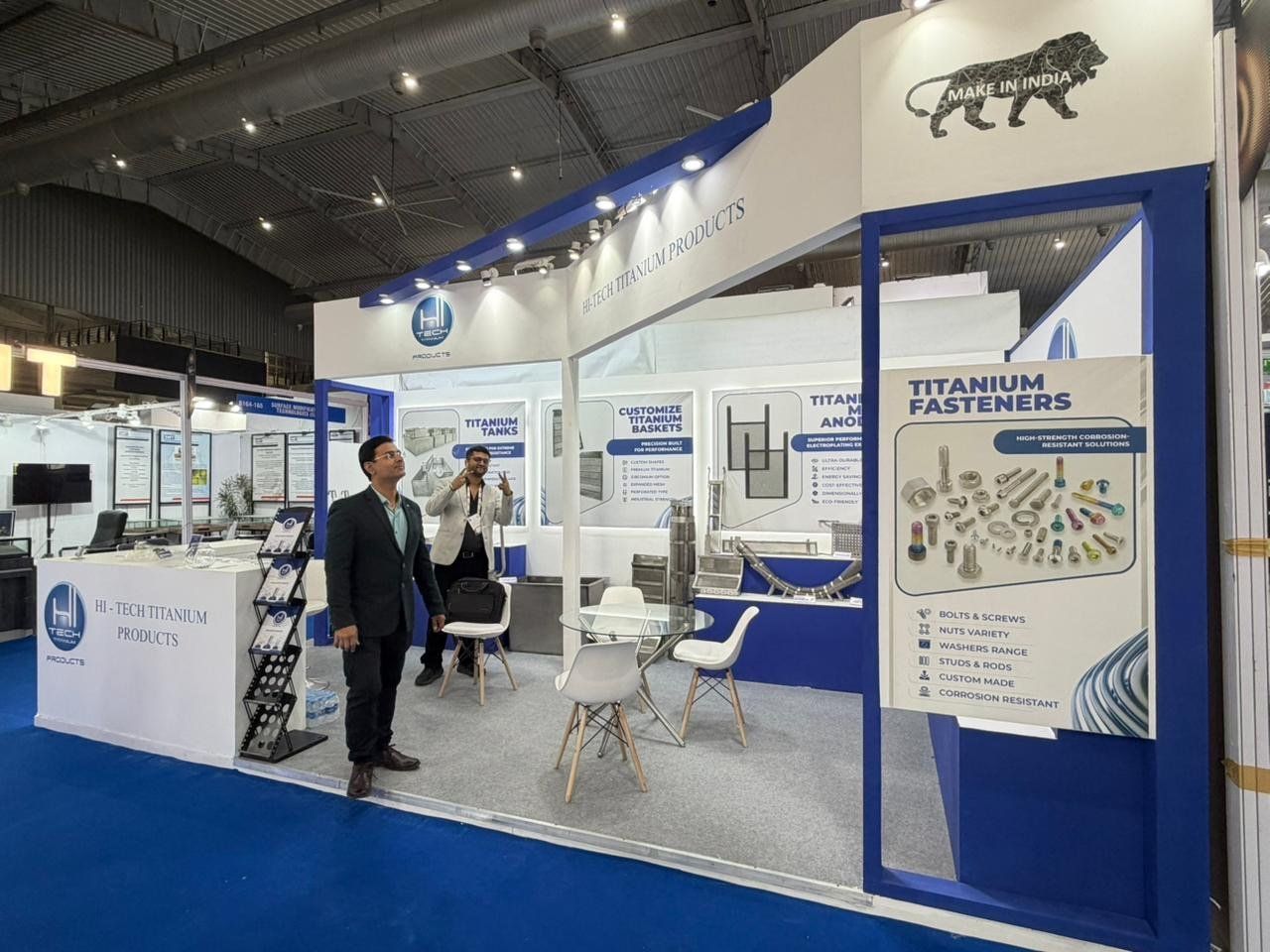 White and blue corner stall with titanium fasteners graphic, product panels and a glass meeting table